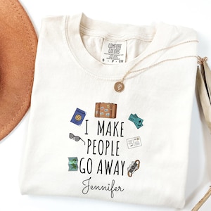 May include: A cream-coloured t-shirt with the text "I MAKE PEOPLE GO AWAY" and a personalised name. The shirt features travel-themed graphics, including a passport, suitcase, sunglasses, and tickets. A gold necklace and a brown hat are also visible.