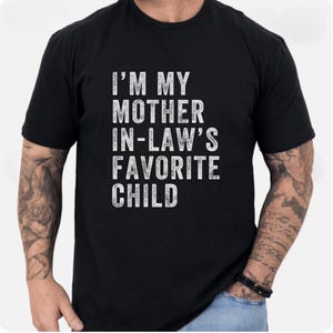Op de afbeelding: Zwart T-shirt met de witte tekst "I'M MY MOTHER IN-LAW'S FAVORITE CHILD". De tekst is in vier regels gestapeld. Het shirt heeft een ronde hals en lijkt van katoen te zijn.