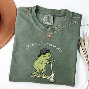 Puede incluir: Camiseta verde oliva Comfort Colors con la frase "off to commit dad foolery" y una rana de dibujos animados montada en un scooter. La rana lleva una gorra de béisbol negra al revés. Un collar dorado y un sombrero marrón también están en la imagen.
