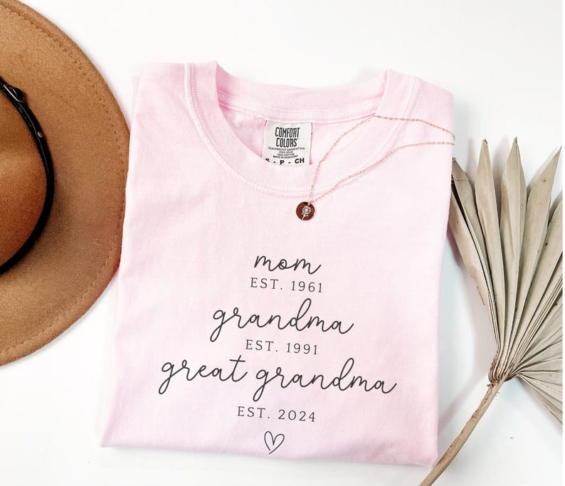 May include: A light pink t-shirt with the words "mom, grandma, great grandma" and the years 1961, 1991, and 2024 printed in black. A gold necklace with a pendant is on the shirt. A brown hat and dried plant are in the background.