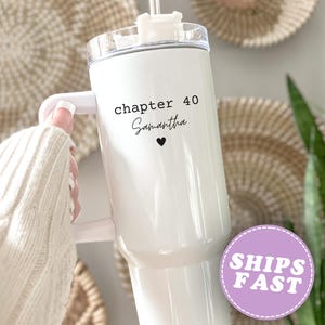 May include: White insulated tumbler with a handle, featuring the text "chapter 40 Samantha" and a heart symbol. The tumbler has a clear lid and a straw. The background includes woven wall decor and greenery.