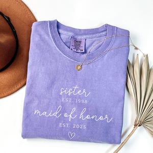 May include: A lavender Comfort Colors t-shirt with the words "sister" and "maid of honor" in white script. The shirt includes the years "EST. 1998" and "EST. 2025" and a small heart. A gold necklace and a brown hat are also in the image.