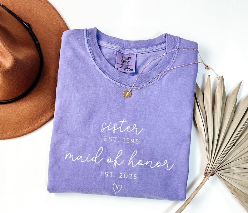 May include: A lavender Comfort Colors t-shirt with the words "sister" and "maid of honor" in white script. The shirt includes the years "EST. 1998" and "EST. 2025" and a small heart. A gold necklace and a brown hat are also in the image.