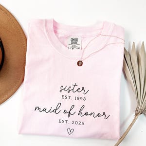 May include: A light pink t-shirt with the words "sister EST. 1998" and "maid of honor EST. 2025" in black script. A gold necklace with a small pendant rests on the shirt. A brown hat and dried plant are also in the image.