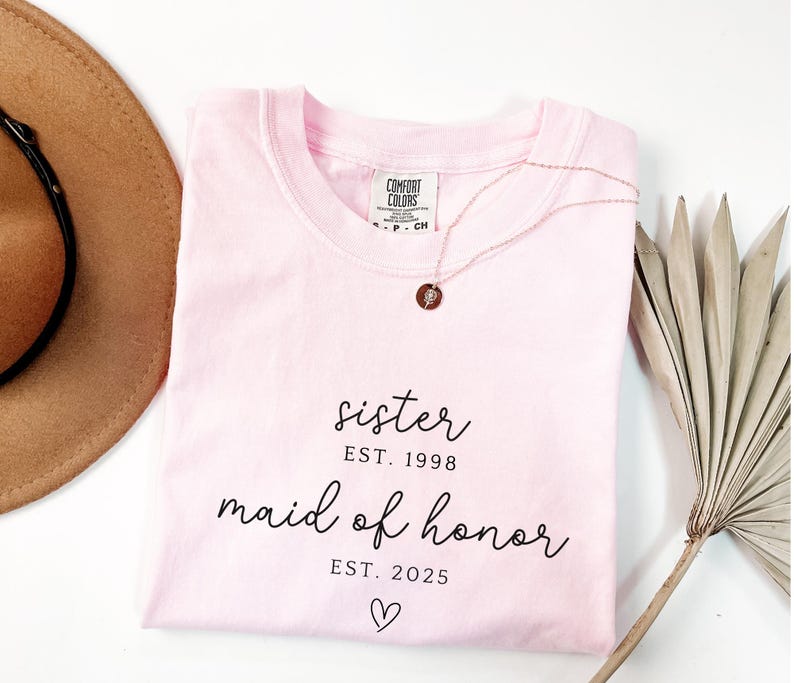 May include: A light pink t-shirt with the words "sister EST. 1998" and "maid of honor EST. 2025" in black script. A gold necklace with a small pendant rests on the shirt. A brown hat and dried plant are also in the image.