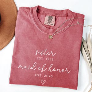 May include: A dusty rose Comfort Colors t-shirt with the words "sister EST. 1998 maid of honor EST. 2025" in white script. A gold necklace with a small pendant rests on the shirt. A brown hat is partially visible.
