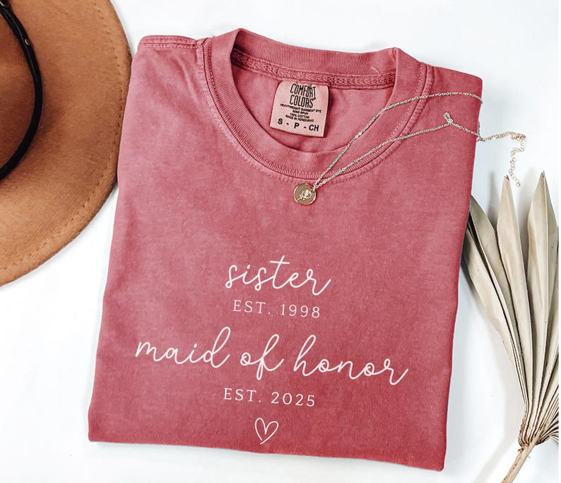 May include: A dusty rose Comfort Colors t-shirt with the words "sister EST. 1998 maid of honor EST. 2025" in white script. A gold necklace with a small pendant rests on the shirt. A brown hat is partially visible.