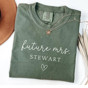 May include: Olive green t-shirt with the words "future mrs. STEWART" in white script, a small heart, and a Comfort Colors tag. A gold necklace with a pendant and a brown hat are also visible.