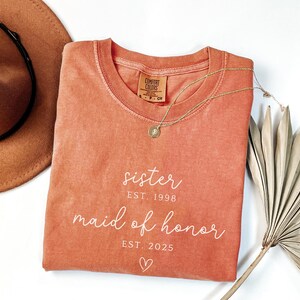 May include: A coral-colored Comfort Colors t-shirt with the words "sister EST. 1998 maid of honor EST. 2025" in white script. A gold necklace and a brown hat are also in the image. A dried palm leaf is in the background.