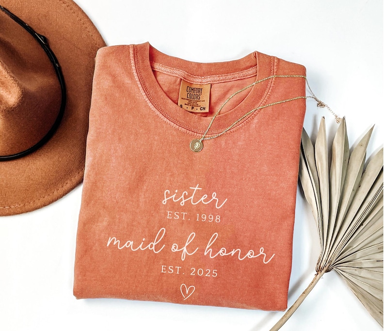 May include: A coral-colored Comfort Colors t-shirt with the words "sister EST. 1998 maid of honor EST. 2025" in white script. A gold necklace and a brown hat are also in the image. A dried palm leaf is in the background.