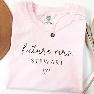 May include: A light pink t-shirt with the words "future mrs. STEWART" in black script, a heart symbol, and a rose gold necklace. The shirt is folded and has a Comfort Colors tag.