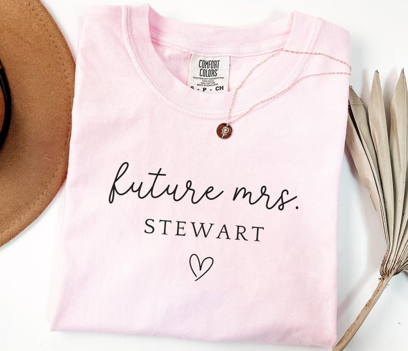 May include: A light pink t-shirt with the words "future mrs. STEWART" in black script, a heart symbol, and a rose gold necklace. The shirt is folded and has a Comfort Colors tag.