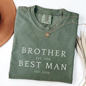 Puede incluir: Una camiseta verde oliva con las palabras "BROTHER BEST MAN" impresas en blanco. La camiseta también tiene los años "EST. 1996" y "EST. 2026" impresos. Un collar dorado con un colgante está sobre la camiseta.