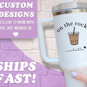 May include: White insulated tumbler with a handle, featuring a graphic of an iced coffee with the words "on the rocks" and a name. The tumbler also has the text "Custom Designs" and "Ships Fast!"