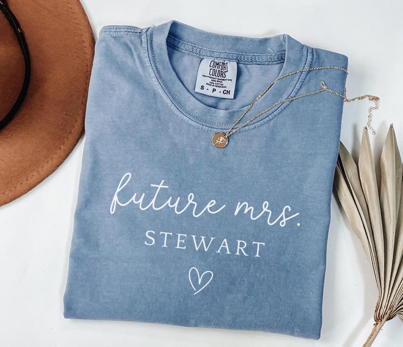 May include: A blue Comfort Colors t-shirt with the words 'future mrs. Stewart' printed in white script. A small white heart is below the name. A gold necklace with a small pendant rests on the shirt. A brown hat is in the upper left corner.