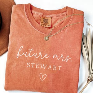 May include: A coral-colored t-shirt with the words "future mrs. STEWART" in white script, along with a small heart. A gold necklace with a pendant rests on the shirt. A brown hat and dried palm leaves are in the background.