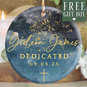May include: Round ceramic ornament with a watercolor design in shades of blue and gold. The ornament features the name "Gideon James", the word "DEDICATED", the date "09.05.25", and a cross. A gold string is attached for hanging.