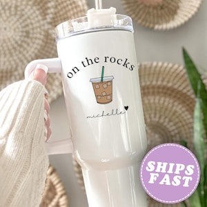 May include: White tumbler with a handle, featuring the text "on the rocks" and a graphic of an iced coffee. The name "michelle" is written below the graphic, with a small heart. The tumbler has a clear lid and a straw.