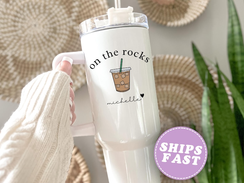 May include: White tumbler with a handle, featuring the text "on the rocks" and a graphic of an iced coffee. The name "michelle" is written below the graphic, with a small heart. The tumbler has a clear lid and a straw.