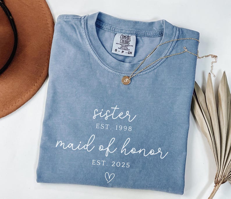 May include: A blue Comfort Colors t-shirt with the words "sister EST. 1998 maid of honor EST. 2025" printed in white. A gold necklace with a pendant is on the shirt. A brown hat and dried plant are in the background.