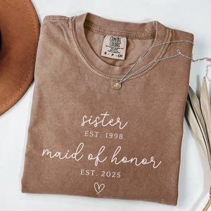 May include: A brown Comfort Colors t-shirt with the words "sister EST. 1998" and "maid of honor EST. 2025" in white script. A silver necklace with a small pendant rests on the shirt. A brown hat is visible in the background.