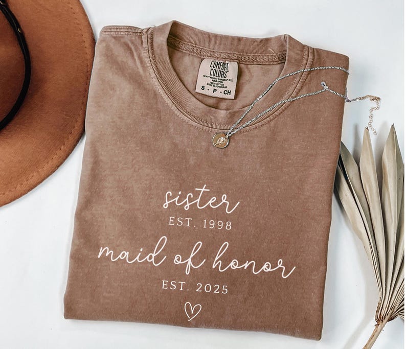 May include: A brown Comfort Colors t-shirt with the words "sister EST. 1998" and "maid of honor EST. 2025" in white script. A silver necklace with a small pendant rests on the shirt. A brown hat is visible in the background.
