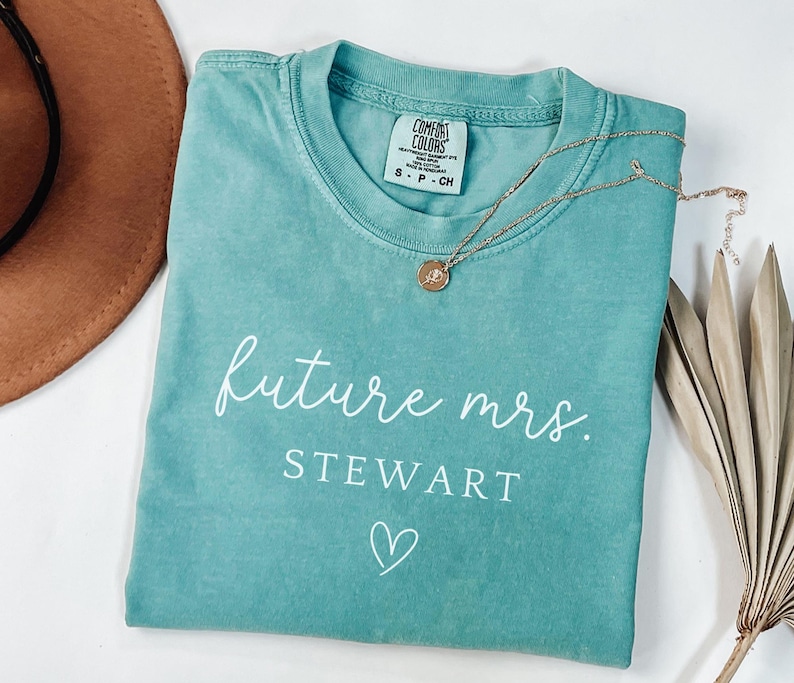 May include: A teal Comfort Colors t-shirt with the text "future mrs. STEWART" and a heart graphic. A gold necklace with a pendant rests on the shirt. A brown hat and dried plant are in the background.