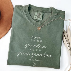 May include: Olive green t-shirt with white text reading "mom EST. 1961, grandma EST. 1991, great grandma EST. 2024" and a heart symbol. A gold necklace with a pendant is draped across the shirt.