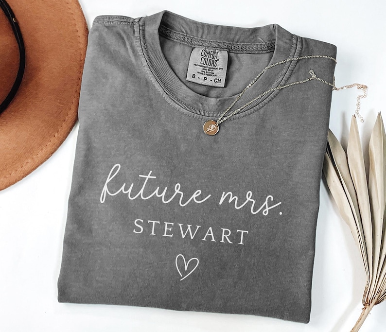 May include: A gray Comfort Colors t-shirt with the words "future mrs. STEWART" in white script, a heart graphic, and a small tag. A gold necklace and a brown hat are also visible.