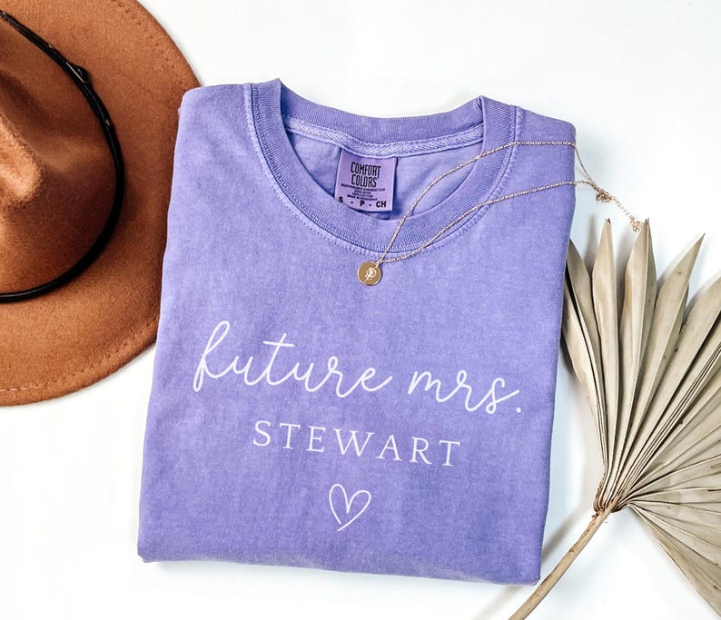 May include: A light purple t-shirt with the words "future mrs. STEWART" in white script, along with a heart symbol. A gold necklace with a pendant rests on the shirt. A brown hat is visible in the background.