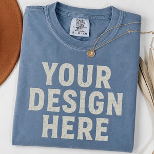 Op de afbeelding: Een blauw t-shirt met korte mouwen, een ronde hals en een Comfort Colors merklabel in de binnenkraag. De voorkant van het shirt is voorzien van de tekst YOUR DESIGN HERE in een verweerd, gebroken wit bloklettertype op de borst.