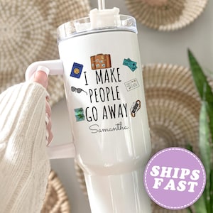 May include: White travel tumbler with a handle, featuring travel-themed stickers and the text "I MAKE PEOPLE GO AWAY" with a name below. The tumbler has a clear lid and a straw. A hand is holding the tumbler.