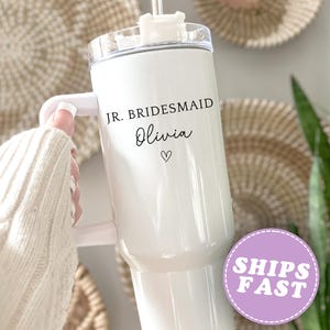 Pu&ograve; includere: Un bicchiere isolante bianco con manico, con la scritta "JR. BRIDESMAID Olivia" e un simbolo a cuore. Il bicchiere ha un coperchio trasparente e una cannuccia. Lo sfondo include decorazioni murali tessute e vegetazione.