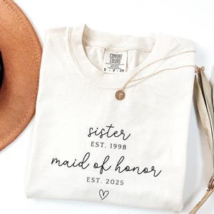 May include: Cream-colored t-shirt with the words "sister EST. 1998 maid of honor EST. 2025" in black script, with a heart symbol. A gold necklace with a pendant rests on the shirt. A brown hat and dried plant are in the background.