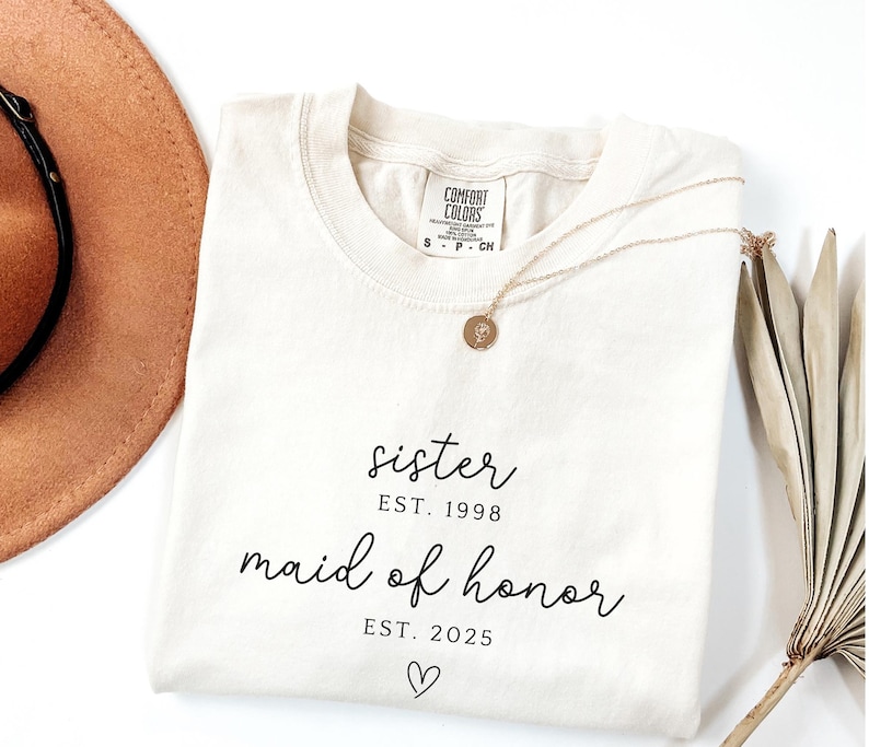 May include: Cream-colored t-shirt with the words "sister EST. 1998 maid of honor EST. 2025" in black script, with a heart symbol. A gold necklace with a pendant rests on the shirt. A brown hat and dried plant are in the background.