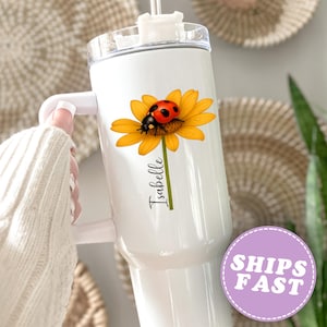 May include: White insulated tumbler featuring a yellow sunflower with a ladybug and the name "Isabelle" in script. The tumbler has a handle and a clear lid with a straw. The design is printed on the side of the tumbler.