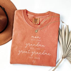 May include: A coral-colored Comfort Colors sweatshirt with the words "mom, grandma, great grandma" and their established years in white script. A gold necklace and a brown hat are next to the shirt. A dried palm leaf is in the background.
