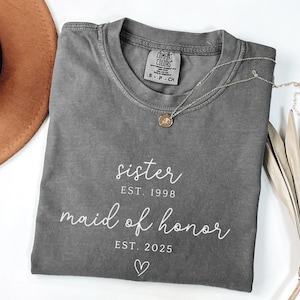 May include: A gray Comfort Colors t-shirt with the words "sister" and "maid of honor" printed in white script. The shirt also has the years "1998" and "2025" printed below the words. A gold necklace with a pendant is on the shirt.