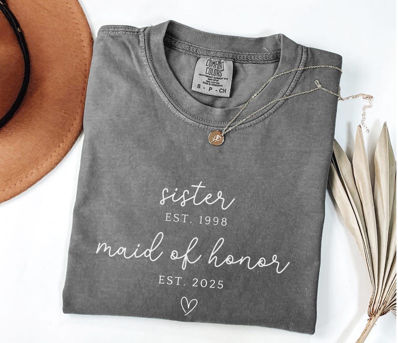 May include: A gray Comfort Colors t-shirt with the words "sister" and "maid of honor" printed in white script. The shirt also has the years "1998" and "2025" printed below the words. A gold necklace with a pendant is on the shirt.