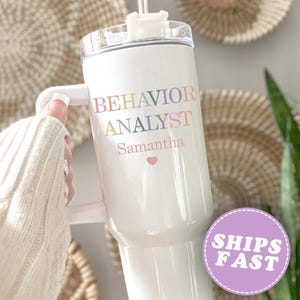 May include: White tumbler with a handle, featuring the words "BEHAVIOR ANALYST" in rainbow colors, the name "Samantha", and a pink heart. A purple sticker with "SHIPS FAST" is visible. The tumbler is held by a person wearing a cream-colored sweater.