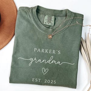 Pode incluir: Uma camiseta verde-oliva com as palavras "Parker's grandma" em escrita branca, um símbolo de coração e "EST. 2025". Um colar de ouro com um pingente repousa sobre a camiseta. Um chapéu marrom é parcialmente visível.