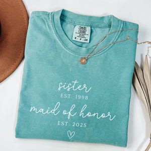 May include: A teal Comfort Colors t-shirt with the words "sister EST. 1998 maid of honor EST. 2025" in white script. A gold necklace with a pendant rests on the shirt. A brown hat and dried plant are in the background.