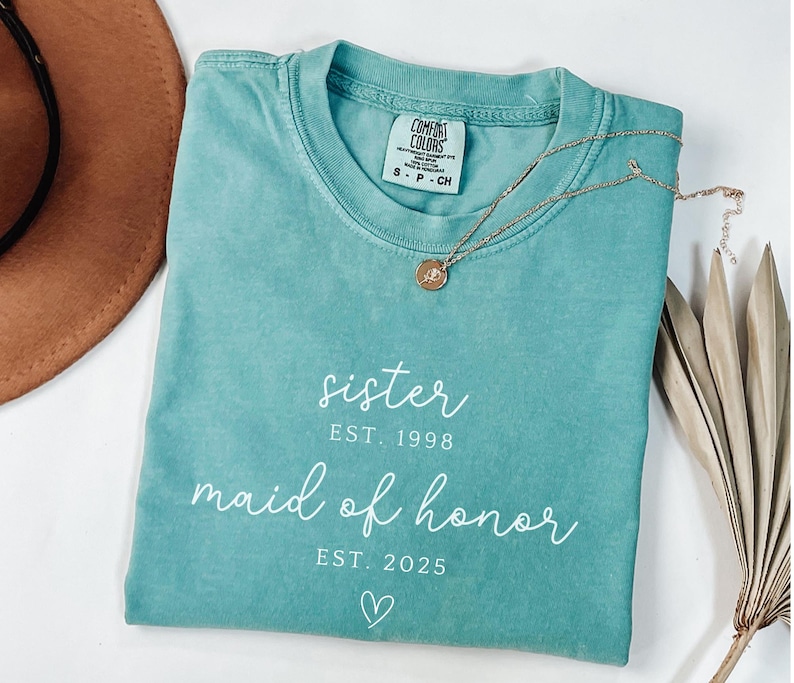 May include: A teal Comfort Colors t-shirt with the words "sister EST. 1998 maid of honor EST. 2025" in white script. A gold necklace with a pendant rests on the shirt. A brown hat and dried plant are in the background.