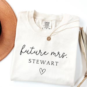 May include: Cream-colored t-shirt with the text "future mrs. STEWART" in black script, a heart symbol, and a gold necklace. A brown hat and dried leaves are in the background. The shirt has a Comfort Colors tag.