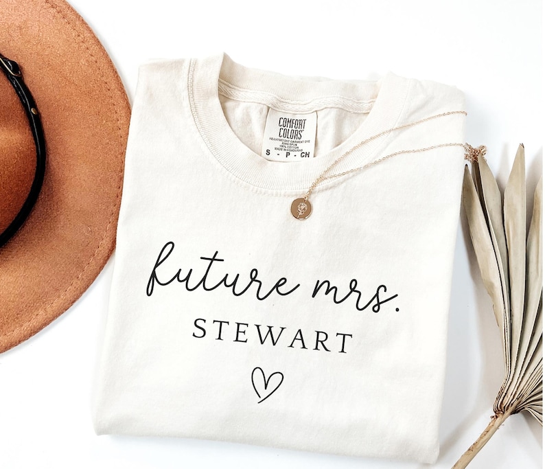May include: Cream-colored t-shirt with the text "future mrs. STEWART" in black script, a heart symbol, and a gold necklace. A brown hat and dried leaves are in the background. The shirt has a Comfort Colors tag.