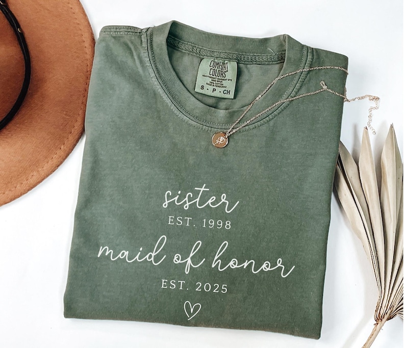 May include: An olive green t-shirt with the words "sister" and "maid of honor" printed in white script. The shirt also has the years "EST. 1998" and "EST. 2025" printed below the words. A gold necklace with a small pendant rests on the shirt.