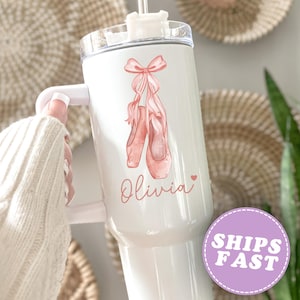 May include: White tumbler with a handle, featuring a watercolor illustration of pink ballet shoes tied with a bow. The name "Olivia" is written in cursive below the shoes, with a small heart. A "SHIPS FAST" sticker is visible.