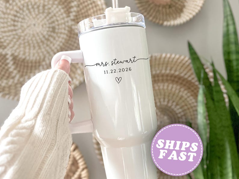 May include: White insulated tumbler with a handle. The tumbler has the text "mrs. stewart", the date "11.22.2026", and a heart symbol. The tumbler has a clear lid and a straw. The background includes woven wall decor and greenery.