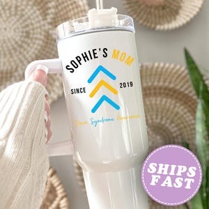 May include: White insulated tumbler with a handle, featuring the text "SOPHIE'S MOM" in black, with blue and yellow arrows, and the words "Down Syndrome Awareness." The year "2019" is also printed on the tumbler.