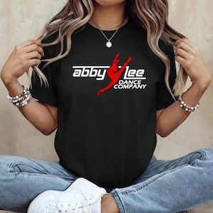 May include: Black t-shirt featuring the text "abby lee DANCE COMPANY" in white and red. The design includes a stylized dancer in red. The shirt is a crew neck style.